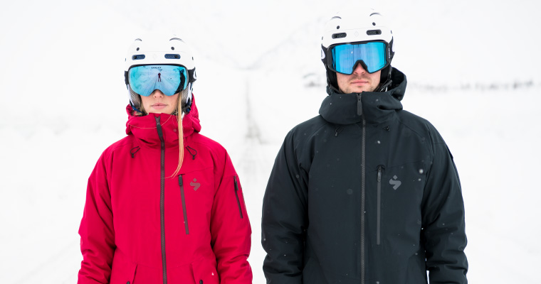 A male and a female skier wearing Sweet Protection goggles, ski helmets, and snow apparel