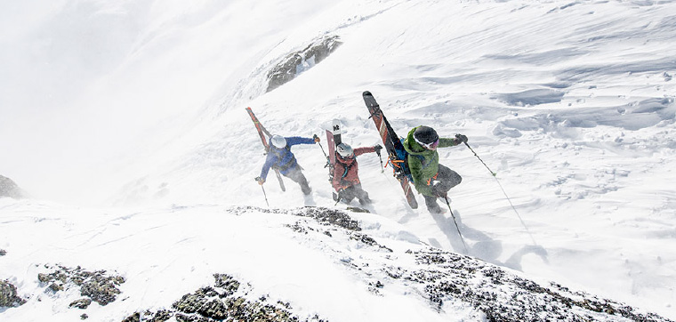 Three skiers bootpack up a steep slope on a windy day
